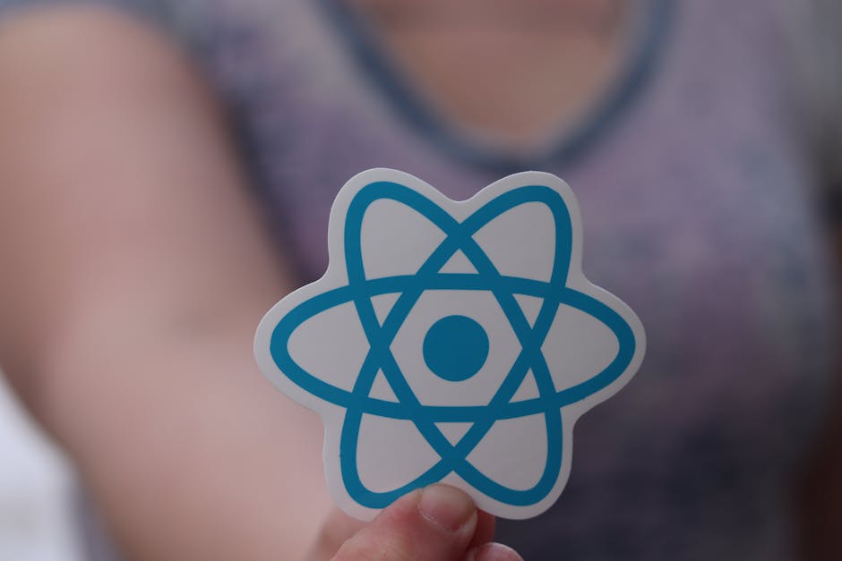Close-up of a person's hand holding a white sticker with a blue atomic model symbol, featuring interconnected orbits and a central dot, in front of a blurred background showing a person wearing a purp