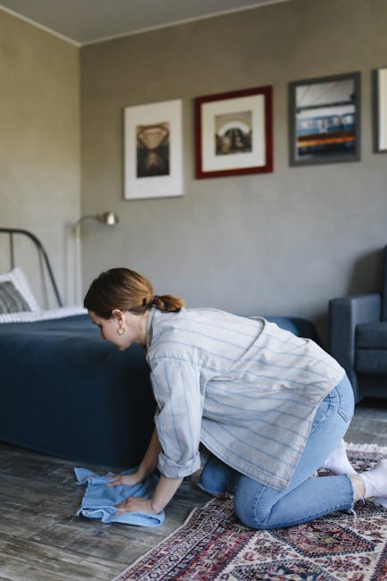 Close-up of a person using a vacuum cleaner with a black hose and silver wand attachment on a light wood laminate floor in a residential room. The individual, dressed in dark shorts and grey slippers,